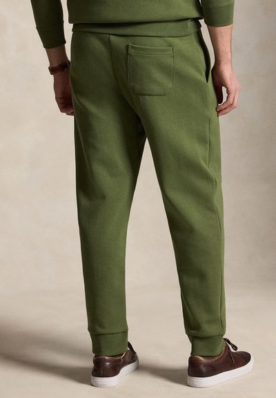 Férfi olívazöld melegítőnadrágban és hozzá illő pulóverben, hátulról látható, sötétbarna tornacipő fehér talppal párosítva.