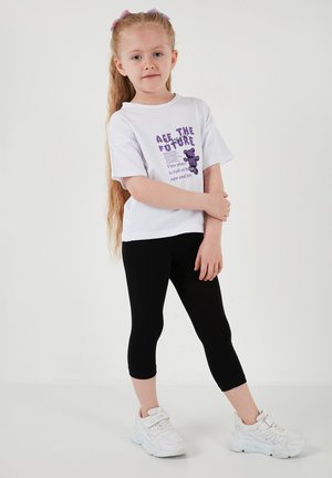 Noor tüdruk pikade juustega, kes kannab valget t-särki lilla tekstiga ja musti lühikesi retuuse, seisab valge ühevärvilise tausta ees.
