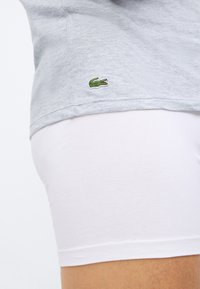 Grå bomull T-skjorte med en grønn brodert krokodillelogo på nederkant, sammen med hvite bomull boxershorts, delvis synlige.