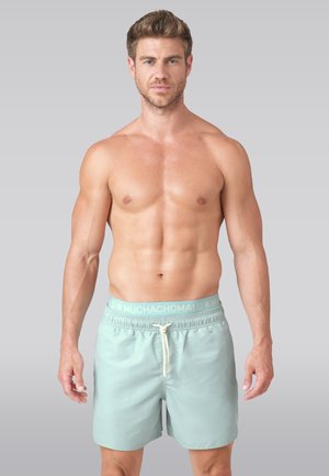 Muž bez trička s krátkymi vlasmi, oblečený v svetlomodré plavky s logom na páse a bielou šnúrkou na jednofarebnom pozadí.