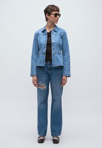 Γυναίκα με κοντά μαλλιά που φοράει γυαλιά ηλίου, ένα ανοιχτό μπλε τζιν μπουφάν, σκισμένα μπλε τζιν και μαύρα φλατ παπούτσια, στέκεται μπροστά σε ένα απλό φόντο.