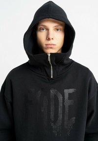 Młoda osoba o niebieskich oczach, ubrana w czarną bluzę z kapturem z podniesionym zamkiem, z przodu wyraźny tekst „FADE” w fakturowanym stylu.