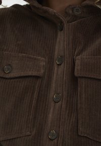 Bruine corduroy shirt met een kraag, twee zakken met klep en donkere knopen. De stof heeft een geribbelde textuur met verticale lijnen.