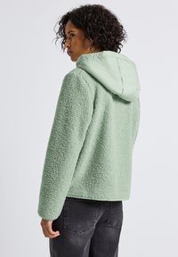 Persona con capelli ricci che indossa una giacca in pile verde chiaro con cappuccio morbido, di spalle e che guarda a sinistra su uno sfondo semplice.