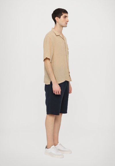 Beige linen short-sleeve shirt, navy shorts, white sneakers with green accents; relaxed fit, soft texture, and button-down front.