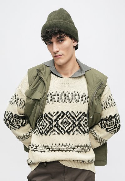 Kötött gyapjú pulóver krém alappal és fekete geometriai mintákkal, olajzöld ujjatlan kabáttal és szürke galléros inggel párosítva.