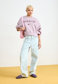 Lila Nike Sweatshirt mit übergroßen Ärmeln, hellblauen weiten Jeans und lila Schuhen, trägt eine rosa Handtasche vor einem neutralen Hintergrund.