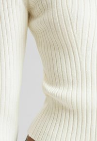 Pull crème côtelé avec rayures verticales, texture douce, coupe ajustée et col en V subtil. Les détails visibles du tissu accentuent la structure.