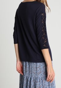 Mörkblå topp med trekvartsärmar i spets och lös passform, kombinerad med en ljusblå blomstermönstrad kjol. Mjuk tyg och texturerade detaljer.