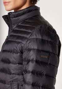 Detailní záběr na osobu v černé prošívané bundě s vysokým límcem a nášivkou loga BOSS na horním rameni.