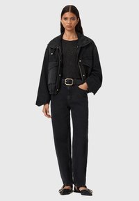 Donna che indossa un giaccone nero oversize, un top nero a pois, jeans neri a vita alta con una cintura larga e ballerine nere appuntite con fibbie.