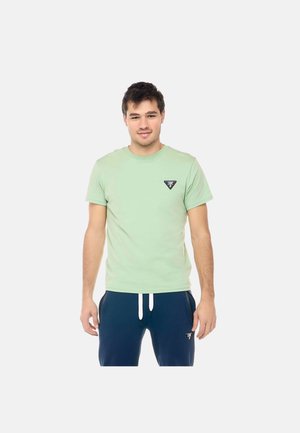 Giovane uomo in piedi che indossa una t-shirt verde chiaro e pantaloni della tuta blu navy con coulisse bianche su uno sfondo chiaro e semplice.