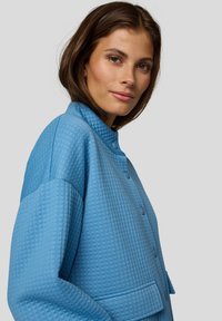 Blue quilted oversized jacket with a mandarin collar, featuring textured fabric, front pockets, and button closure.