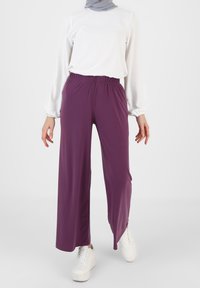 Paarse wijde broek gemaakt van zachte stof met een elastische tailleband, gecombineerd met een witte lange mouwen top en witte sneakers.