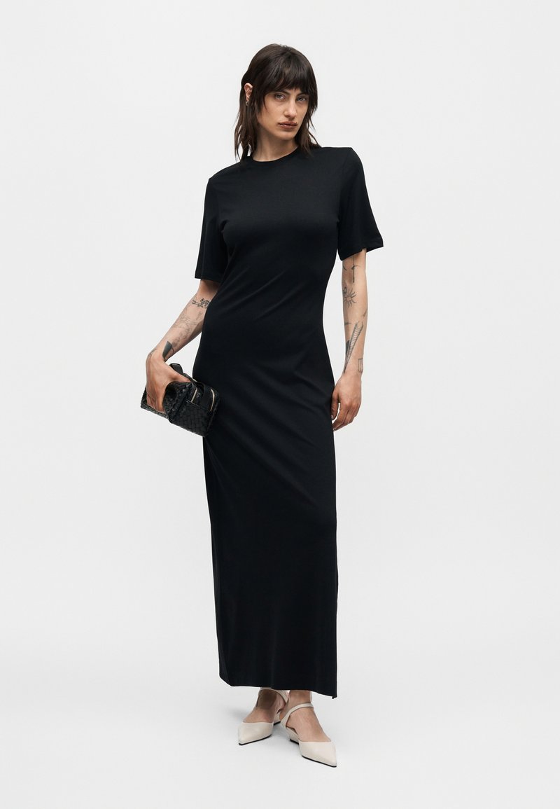 Frau mit dunklem Haar und Tattoos, die ein schwarzes Maxikleid mit kurzen Ärmeln trägt, eine schwarze Clutch hält und weiße spitze Slingback-Schuhe trägt.