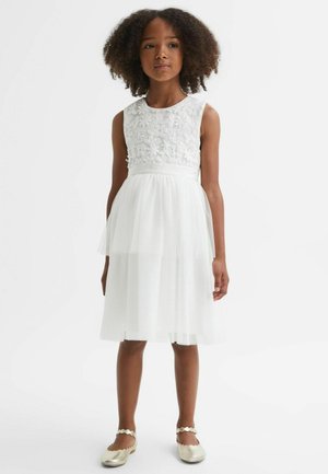 Młoda dziewczyna z kręconymi włosami, ubrana w białą sukienkę bez rękawów z kwiatowym haftem i przezroczystą, warstwową spódnicą, zestawioną ze złotymi balerinami.