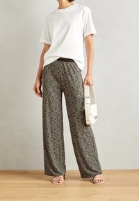 T-shirt blanc avec texte en relief et pantalon large noir à motif abstrait blanc. Le modèle tient un sac blanc et porte des sandales.