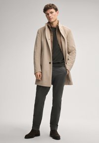 Beiger Wollmantel mit Reverskragen und Knopfverschluss über einem dunklen Pullover. Das Model trägt dunkelgrüne Hosen und braune Wildleder-Stiefel.