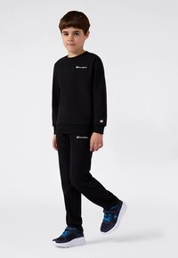 Ragazzo che indossa una felpa e pantaloni neri Champion, con sneakers blu e bianche, in piedi con un piede leggermente sollevato su uno sfondo bianco.