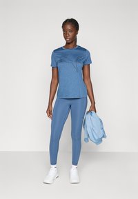 Blå atletisk t-shirt med en svart outline-logotyp, parat med matchande blå leggings. En ljusblå jacka hålls i vänster hand.