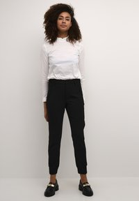 Top bianco a maniche lunghe con scollo arricciato, abbinato a pantaloni neri a sigaretta e mocassini neri con dettagli in metallo dorato.