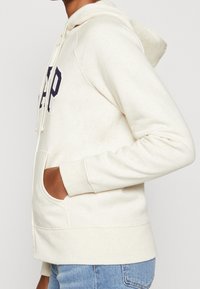 Haut de survêtement crème clair avec une poche kangourou, capuchon à cordon de serrage et lettrage "DEP" en marine. Texture douce et coupe décontractée.