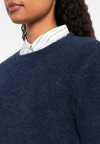 Egy nő alsó arca és nyaka látható, sötétkék, bolyhos pulóvert visel, amely fehér ing fölé van rétegezve, amelyen vékony fekete függőleges csíkok találhatók.