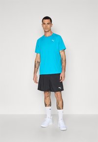 Turkis atletisk t-shirt med korte ærmer, logo på brystet, kombineret med sorte shorts og hvide sneakers. Glat stofstruktur.