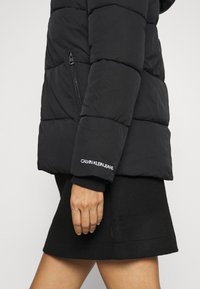 Czarna kurtka puchowa o pikowanej strukturze, z zamkiem błyskawicznym w kieszeni i haftowanym logo "Calvin Klein Jeans" na mankiecie. Prosty, funkcjonalny design.