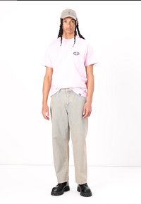 Camiseta rosa claro con un gráfico redondo en el lado izquierdo del pecho, combinada con jeans beige holgados y deslavados y botas negras con suela gruesa. Gorra gris.