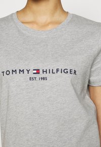 Šedé bavlněné tričko s nápisem "TOMMY HILFIGER" v námořnické modři, s malým logem v červené, bílé a modré nad nápisem "EST. 1985" ve středu.
