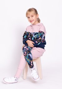 Różowa i granatowa bluza z kapturem w kwiatowy wzór, zestawiona z różowymi spodniami. Modelka ma na sobie białe tenisówki i wzorzyste skarpety, siedząc na stołku.