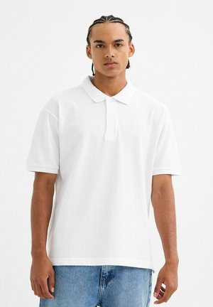 Polo blanco de algodón, con cuello clásico, mangas cortas y tapeta de tres botones. Combinado con jeans de color azul claro.