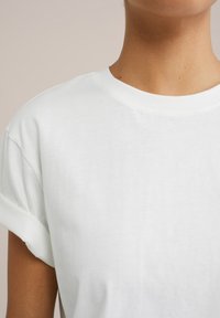 T-shirt en coton blanc avec un col rond et des manches courtes roulées, présentant une texture lisse et un design simple.