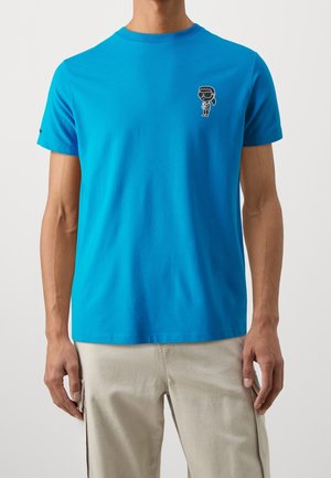 T-shirt en coton bleu à manches courtes, col rond et un petit design graphique noir sur le côté gauche de la poitrine. Texture lisse, coupe classique.