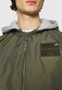 Olivgrüne Bomberjacke mit grauer Kapuze, Reißverschluss vorne und gesticktem Patch mit "Alpha Industries 1959". Glatte, glänzende Oberfläche.