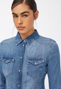 Cămașă din denim de culoare albastră deschisă, cu guler, două buzunare la piept și un accent decorativ cu fundiță din strasuri pe buzunarul din stânga.