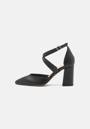 Zapato de tacón alto de cuero negro con punta puntiaguda y tiras en forma de cruz en el tobillo, aseguradas por una pequeña hebilla redonda de plata.