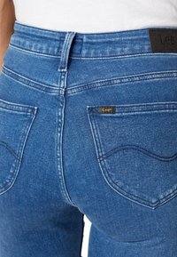 Blå denimjeans med hög midja, med böjda bakfickor och en liten svart etikett på midjebandet. Slät textur hela vägen.