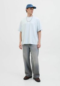 Hellblaustes, übergroßes T-Shirt mit Grafikdruck, kombiniert mit hellgrauen, weiten Jeans und dunkelbraunen Schuhen. Accessoires mit einer blauen Mütze.