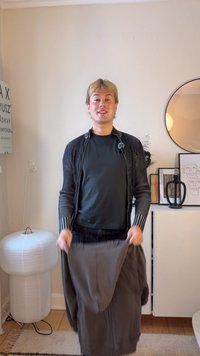 Cardigan à rayures noires et blanches en tricot avec une fermeture éclair, associé à un haut ample gris foncé. Tenant un pantalon gris, debout à l'intérieur près de la décoration.