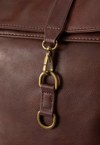 Detalle de una bolsa de cuero marrón con un clip de latón y un lazo de sujeción, con costuras visibles y una superficie texturizada que añade profundidad.
