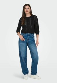 Crna pletena kardigan jakna s kopčanjem na dugme i otvorenim uzorkom, u kombinaciji s labavim plavim trapericama i bijelim tenisicama.