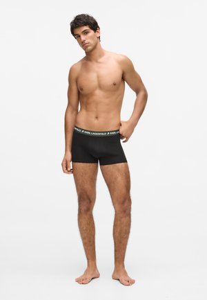 Jauns vīrietis stāv basām kājām, bez krekla, valkājot melnas Karl Lagerfeld bokserbikses, ar vienu roku novietotu pie jostasvietas.