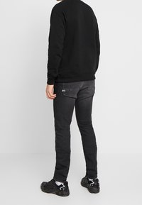 Persona de pie, de espaldas, vistiendo un sudadera negra, jeans ajustados gris oscuro con bolsillos traseros, calcetines blancos y zapatillas negras sobre un suelo blanco.