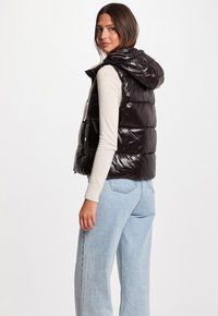 Gilet imbottito nero lucido con cappuccio, caratterizzato da motivi a cucitura orizzontale, tasche con zip e dettagli con logo sulla tracolla. Abbinato a jeans chiari.