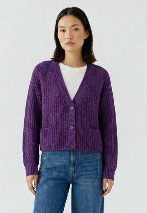 Femme portant un cardigan violet côtelé boutonné par-dessus une chemise blanche et un jean bleu, debout devant un fond clair uni.