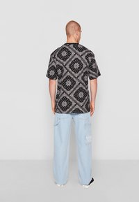 Maglietta nera con motivi geometrici bianchi, maniche corte, indossata con jeans ampi blu chiaro, vista da dietro. Sneakers nere.