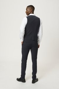 Mornarski suknjični brezrokavnik s prilagojeno zasnovo, nošen čez belo srajco. Hlače so slim-fit z gladko teksturo, dopolnjene z črnimi čevlji.
