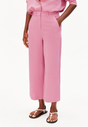 Frau, die hoch taillierte, weit geschnittene rosa Hosen mit Vordertaschen und schwarze Sandalen mit weißen Riemen trägt, steht mit einer Hand in der Tasche.